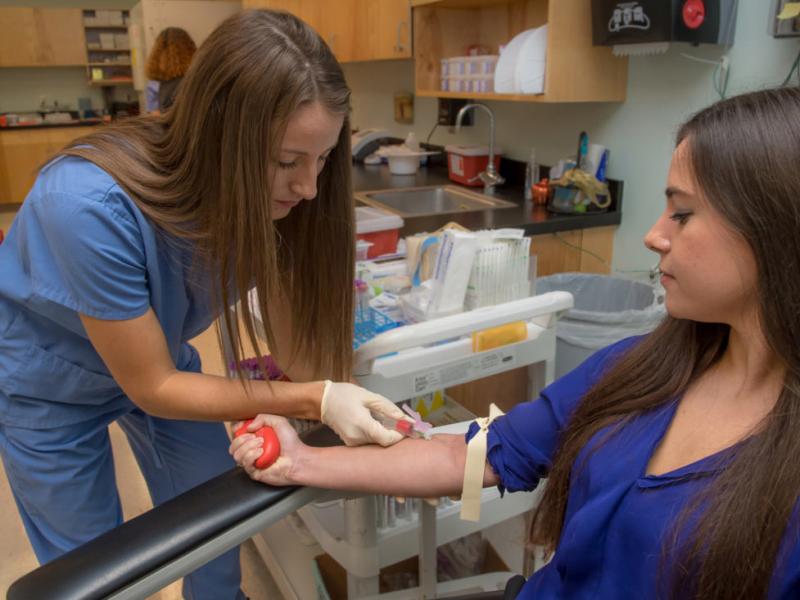 Researcher takes blood sample from another person in a clinical setting.