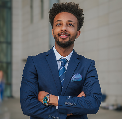 Caleb Hailemariam headshot, standing with arms crossed, wearing a suit, looking into distance. 