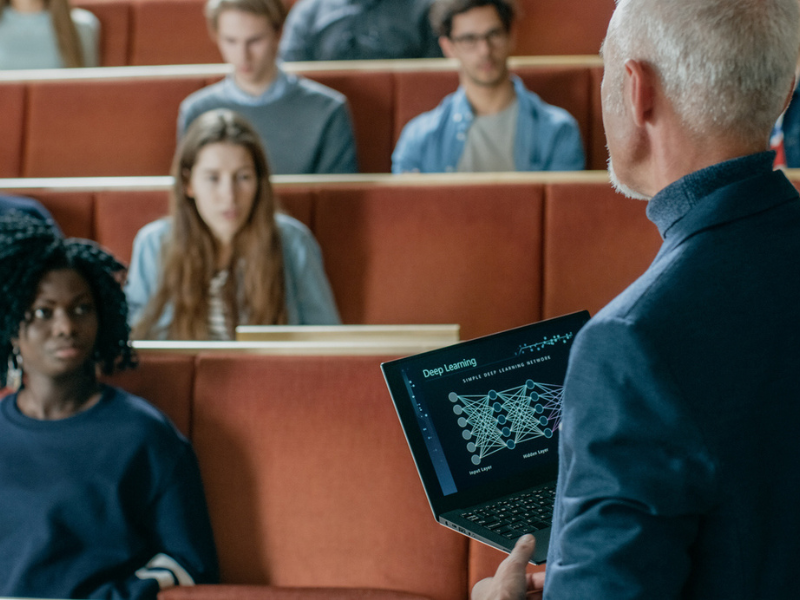 Professor lectures attentive students in a tiered classroom, holding a laptop displaying a graph.