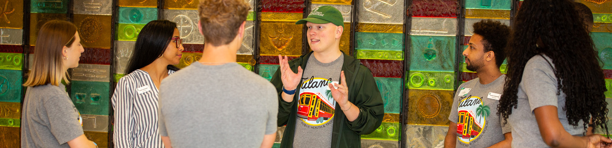Man in green cap talks to diverse students against a colorful mosaic wall.