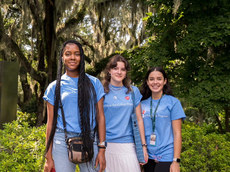 Three smiling young women in blue shirts stand outdoors among green trees.