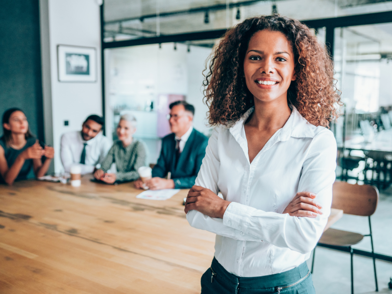 A Black Woman smiles in front of a table, where her work team sits.