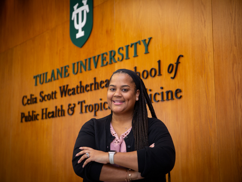 Smiling Leshawn Simplis-Barnes with braids, arms crossed, in front of Tulane Public Health sign.