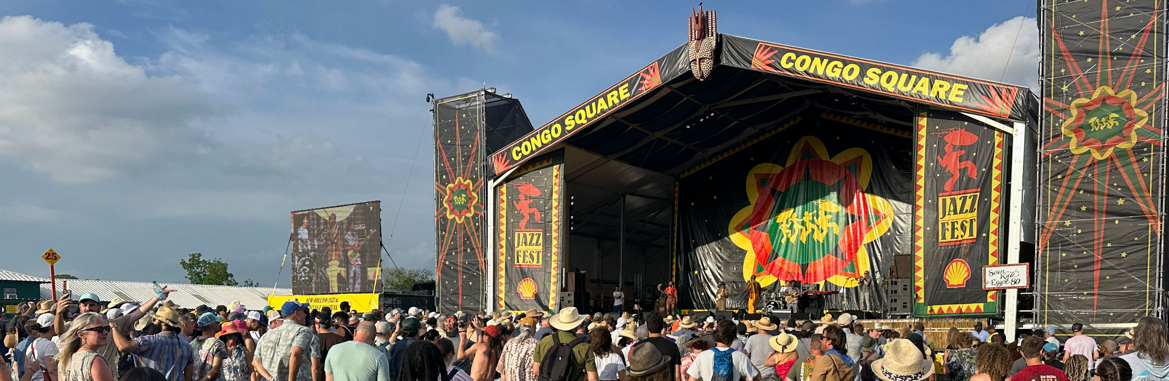 Crowd gathered in front of a large outdoor stage with colorful decorations under a cloudy sky.