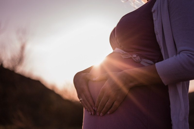 Pregnant person's hands form heart over baby bump at sunset.