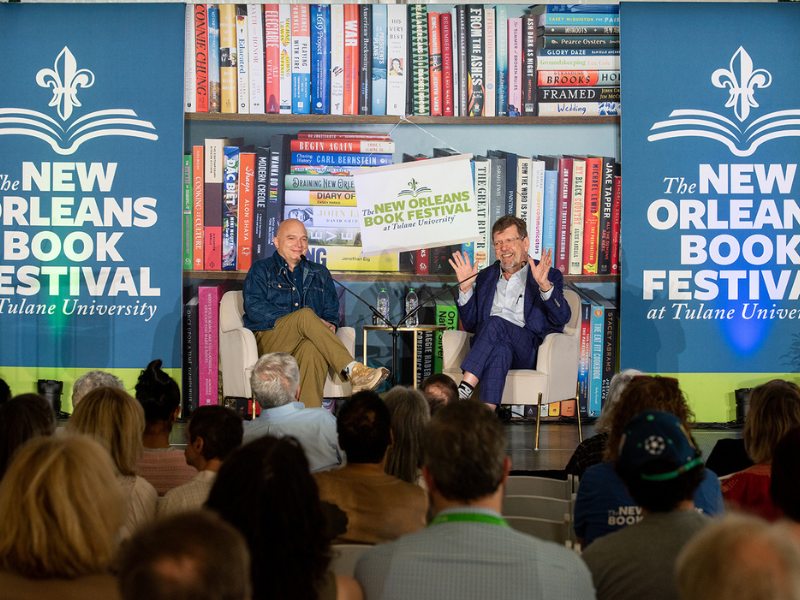 Two men converse on stage at the New Orleans Book Festival, audience visible.