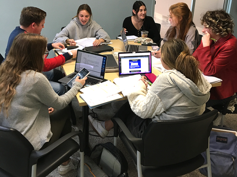 Students collaborate around a table with laptops and books, some smiling.