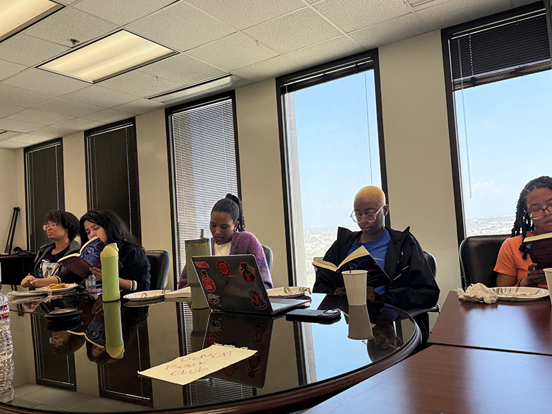 Diverse people focused on reading and laptops around a conference table with large windows.