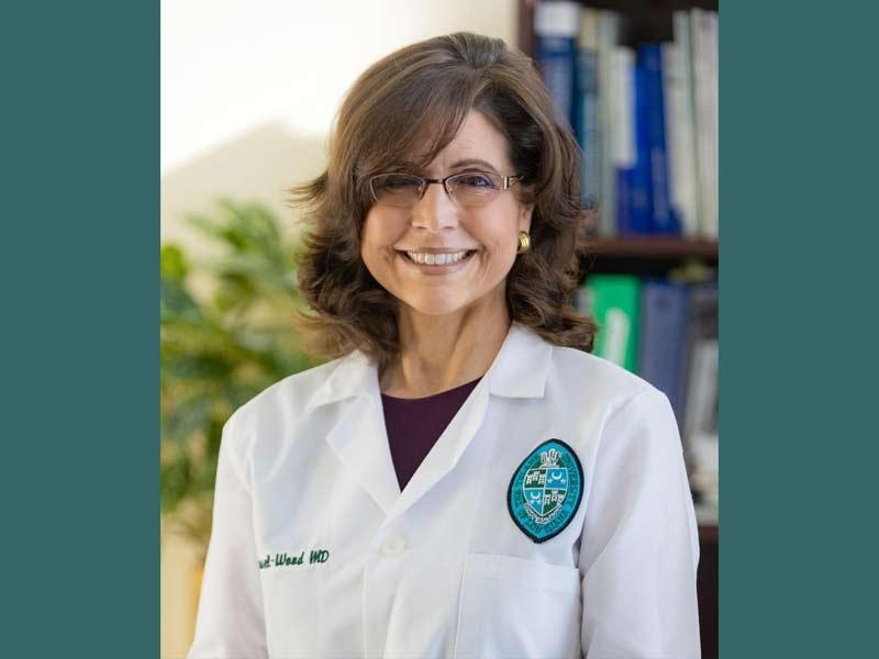 Smiling woman with glasses and brown hair wearing a white lab coat with an emblem.