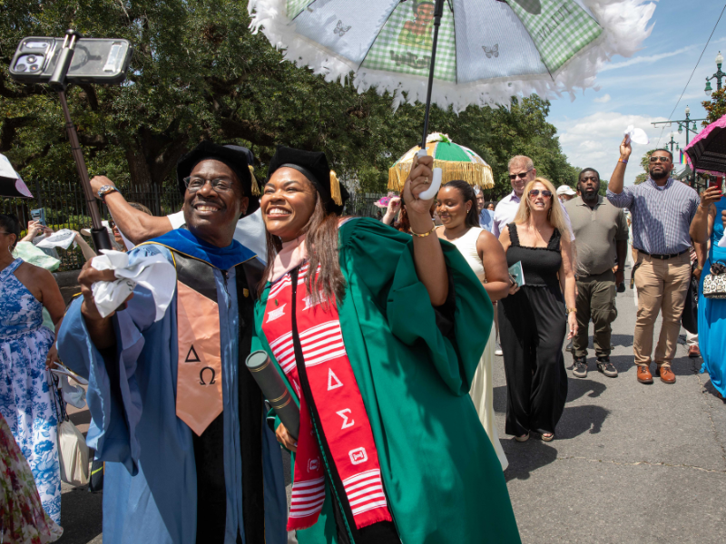 Smiling graduate in green gown takes a selfie with man in blue gown during outdoor celebration.