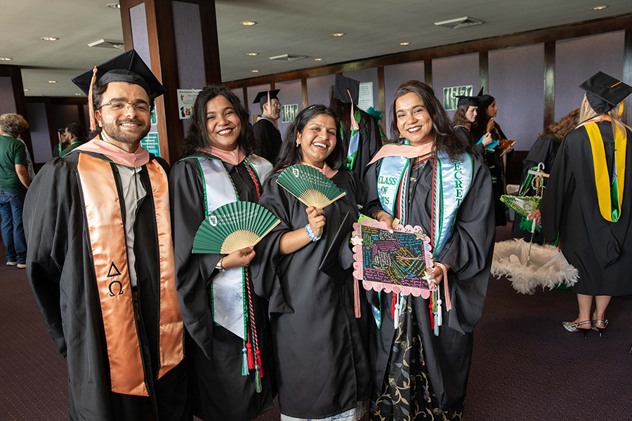 Four smiling graduates in caps and gowns, holding fans and a decorated frame.