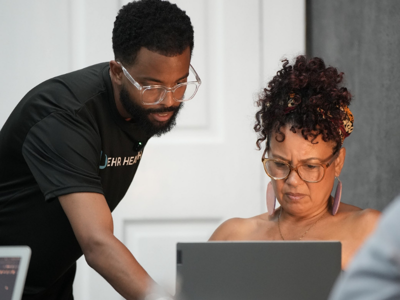 Man leaning over woman's shoulder, both intently looking at a laptop screen.
