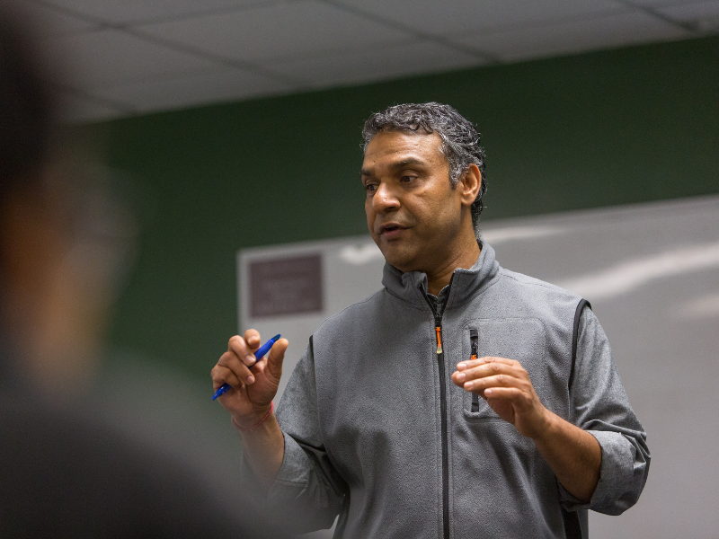 Dr. Sudesh Srivastav speaks, holding a blue pen, gesturing in a classroom.