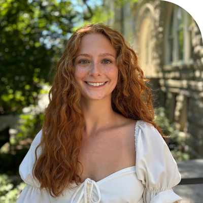 Smiling woman with long red hair wearing a white top, outdoors with stone archway.