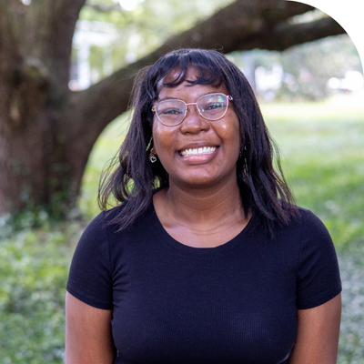 Smiling young Black woman with glasses, bangs, and dark hair, outdoors.