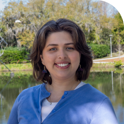 Smiling woman with dark hair and a blue cardigan stands by water and trees.