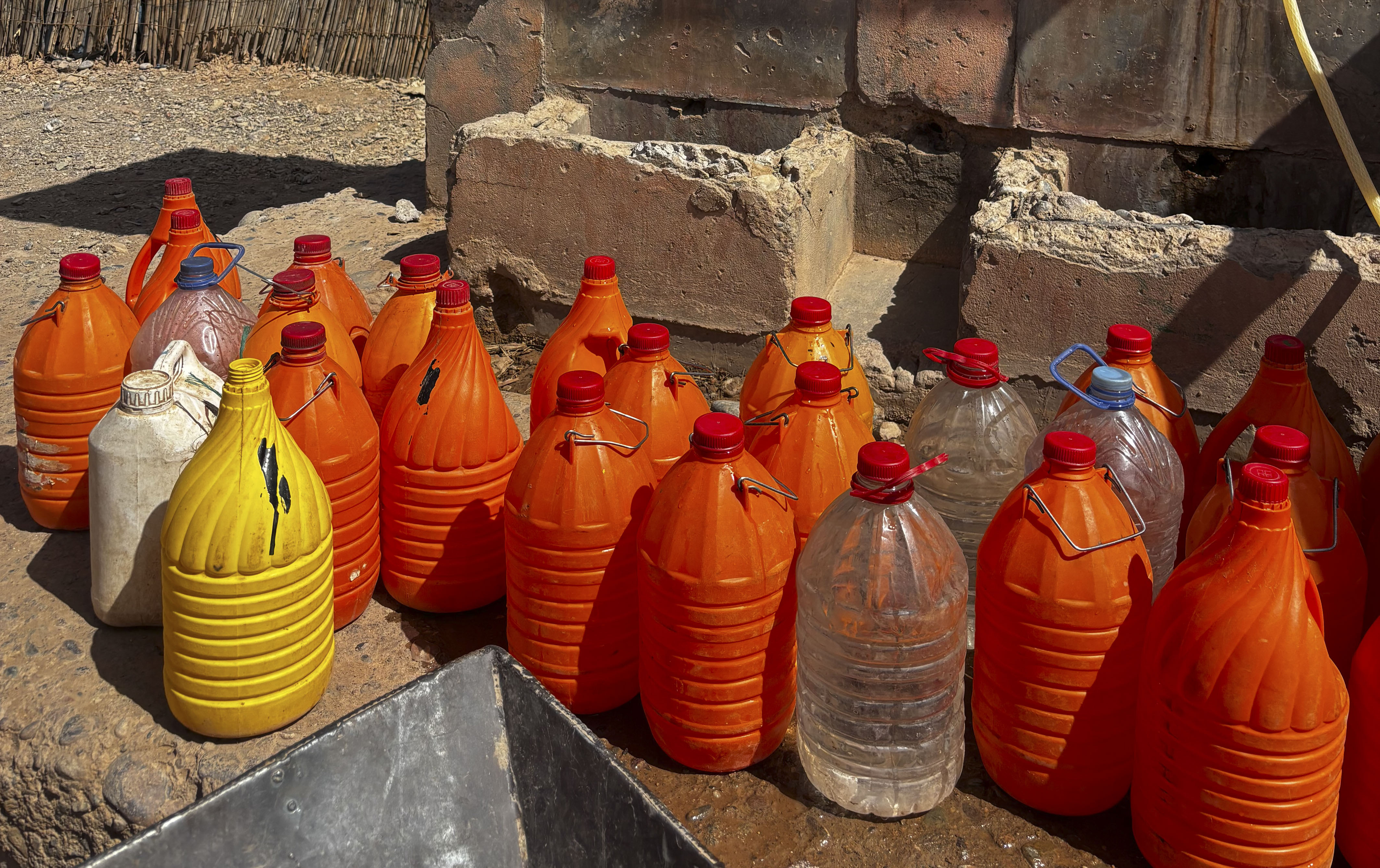 Many orange, clear, and yellow plastic jugs filled with water on dirt ground.