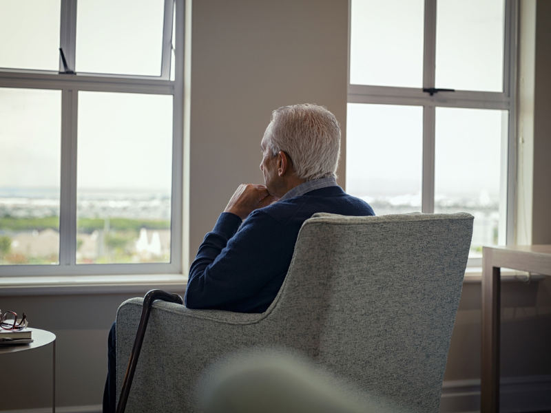 Elderly man with white hair sits with a cane, looking thoughtfully out a large window.
