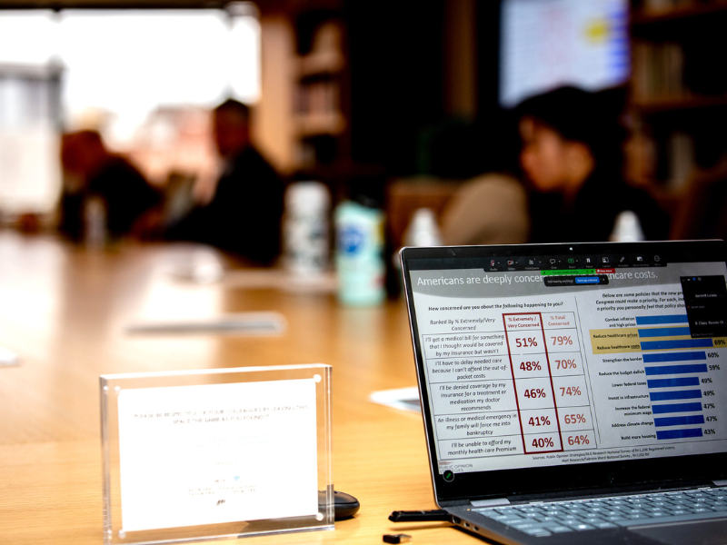 Laptop with data charts on a wooden table; blurred people and bookshelves in background.