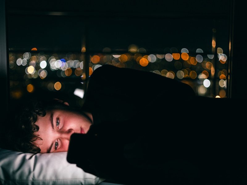 Young person with curly hair lying in the dark staring at phone, bright city lights blurred in background.