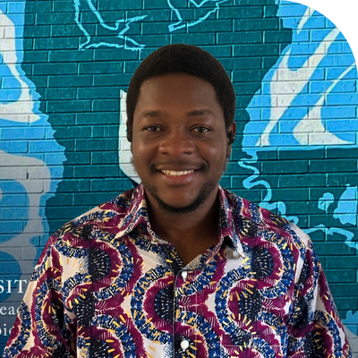 Smiling man in a colorful patterned shirt, standing against a blue brick wall with graffiti.