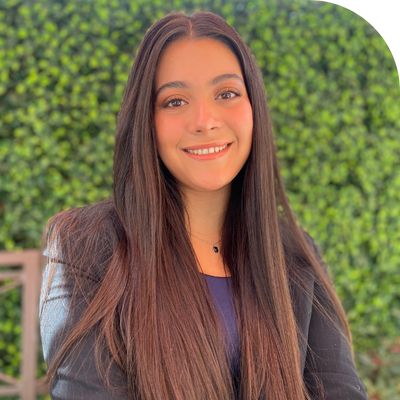 Smiling young woman with long dark hair against a green leafy background.