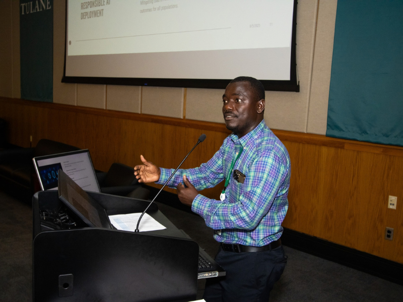 Samuel Kakraba in plaid shirt gestures while speaking at a podium with a presentation screen behind him.