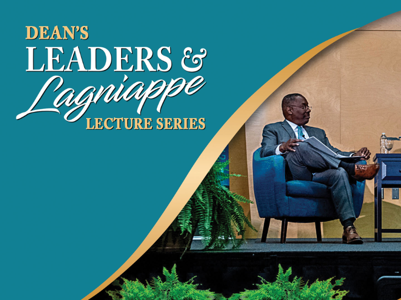 Thomas LaVeist in suit seated on stage, beside a teal and gold lecture series banner.