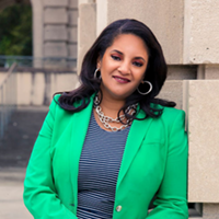 Ashley Shelton, wearing a green suit, smiling, leaning against a building