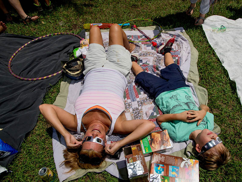 Mother and son viewing eclipse  with glasses, laying on blanket at park