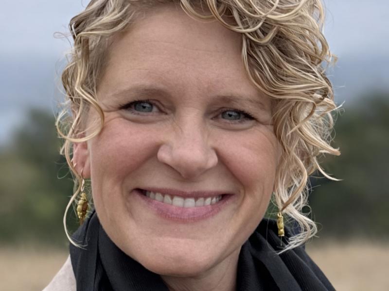Jennifer Makelarski, smiling, with curly blonde hair pulled back, wearing a black scarf, with trees and sky in the background