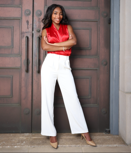Bryson Reems, standing, arms crossed in front of a set of wooden doors, wearing a red blouse and white pants, looking directly at camera