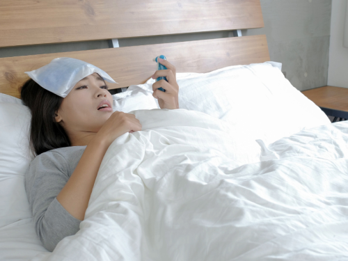 Woman lying in bed with an ice pack on her forehead, holding a phone.