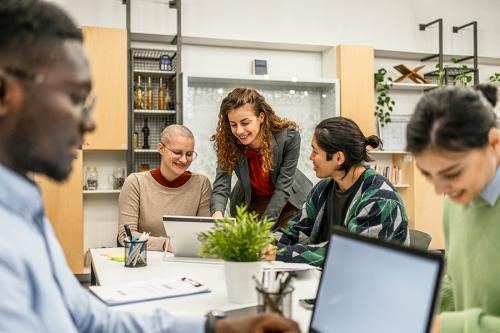 Five coworkers collaborate around a table with laptops in a modern office.