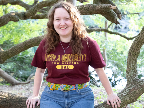 Maeve Karnes, smiling, wearing a Loyloa University t-shirt, sitting on large tree branch. 