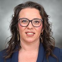 Nichole Valenzuela headshot. Nichole is wearing glasses, smiling, and has long wavy brown hair. 