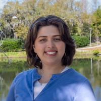 Chloe Garret, smiling,  in blue cardigan outdoors by a lake, trees in background.