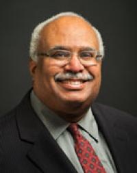 Georges Benjamin, smiling, with dark skin, gray hair, glasses, dark suit, and patterned red tie.
