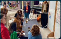 Diverse group of people discussing around a table with a whiteboard in a vibrant room.