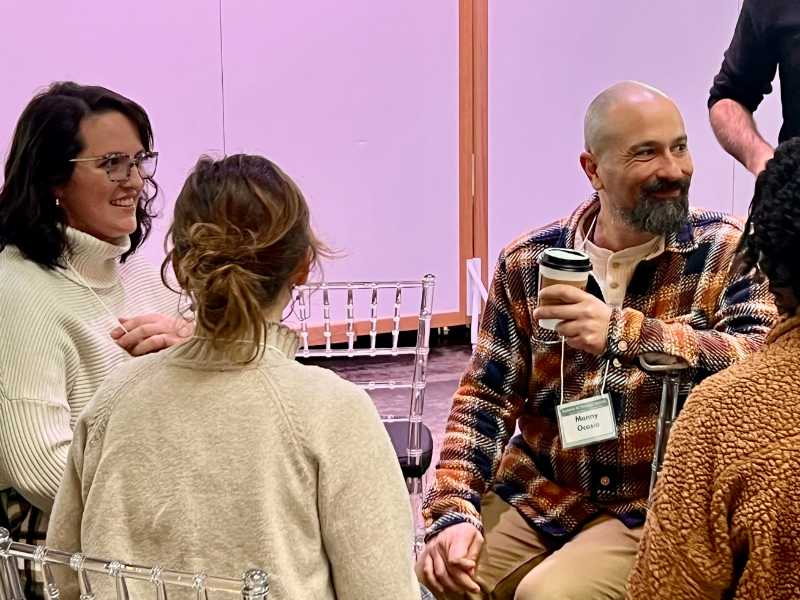 Bearded man with coffee smiles, conversing with two women at an indoor event.