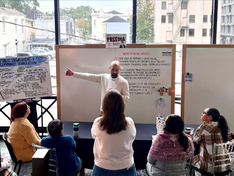 Man in white sweater presents to a small group in a bright room with city views.