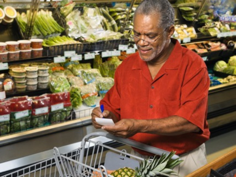 Older Black man checking a shopping list in a grocery store produce aisle.