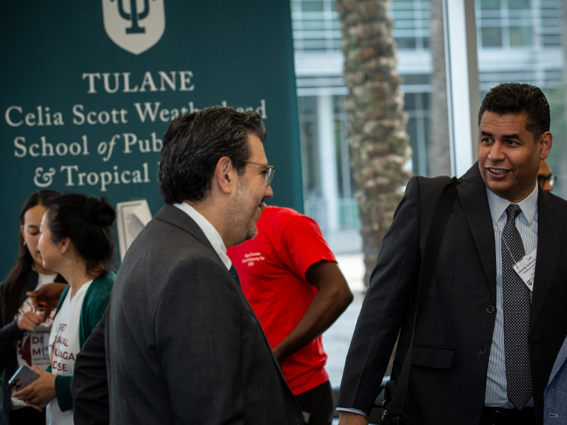 Two men in suits chat at an event; Tulane University banner in background.