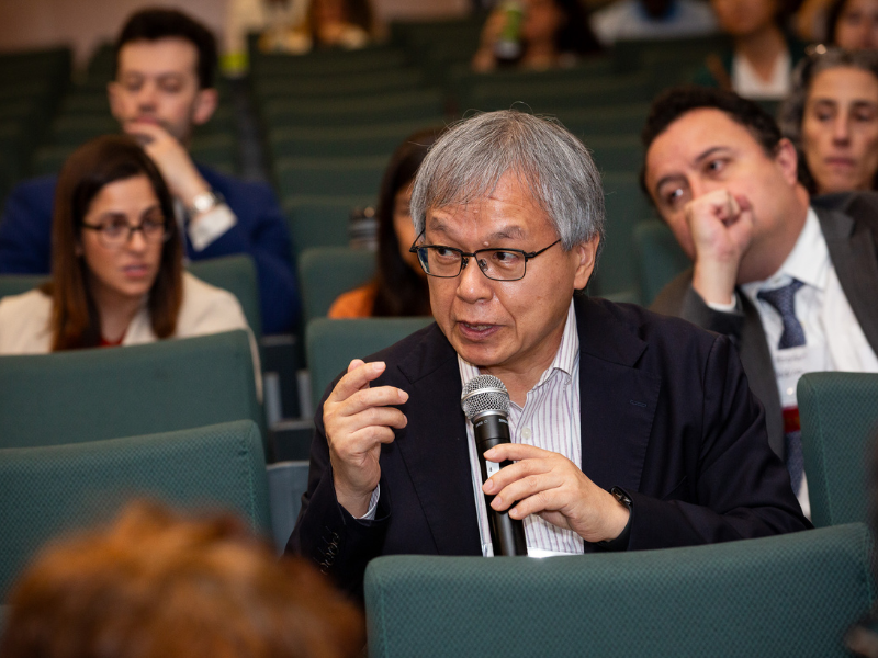 Man with glasses speaking into a microphone in an auditorium, people in background.