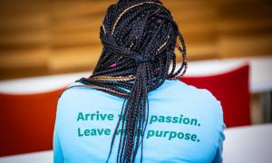 Person with braided hair from behind, wearing a blue shirt with green text: "Arrive with passion. Leave with purpose."