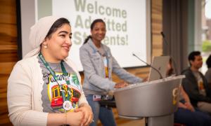 Two smiling women at a student welcome event, one at a podium, the other in the foreground.