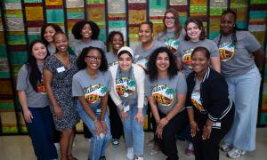 Thirteen smiling diverse women in grey t-shirts with a logo, standing before a colorful mosaic wall.
