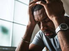 Man holds head in hands with furrowed brow while sitting at table by window. He looks stressed.