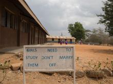 In Photo a sign reads "Girls need to study, don't marry them off." In the background girls are going to a school. 