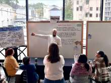 Man in white sweater presents to a small group in a bright room with city views.